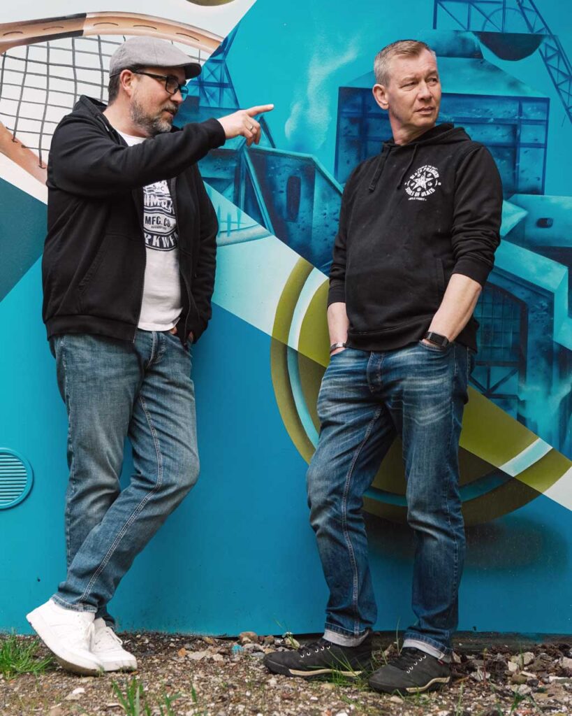 Bandfoto von den Stadtrandpoeten – einem Folk/Pop Duo aus Bochum im Ruhrgebiet. Die Musiker Peter Albert und Oliver Wengeler stehen vor einem blauen Ruhrpott-Graffiti mit Förderturm